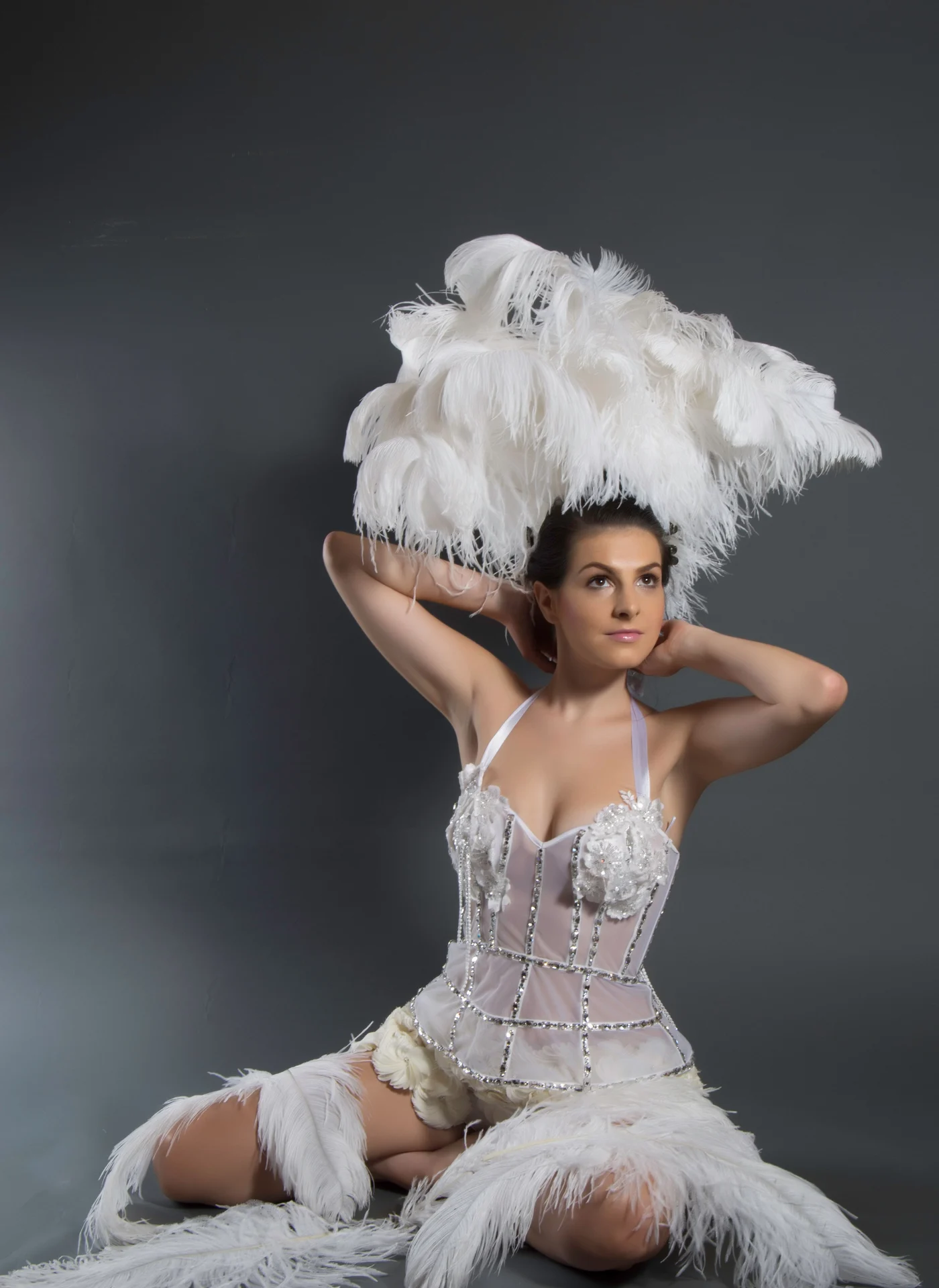 A performer in a towering white ostrich-feather headdress and beaded corset against a dark backdrop